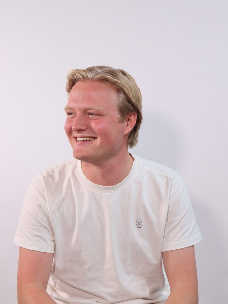 Portretfoto van Roel Vos, een jonge man met blond haar, lachend en gekleed in een wit T-shirt, kijkend naar links tegen een lichte achtergrond.